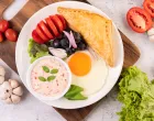 Breakfast consists of bread, fried egg, salad dressing, black grapes, tomatoes, and sliced ​​onions./Freepik