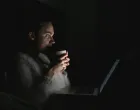 Djevojka leži u krevetu noću i pažljivo gleda u ekran laptopa. Jede hranu, drži šolju i gleda seriju noću./Anastasiia Nelen